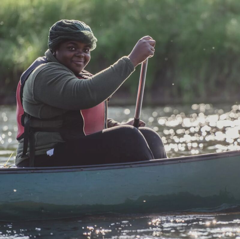 A person is paddling a canoe on a body of water. The individual is wearing a life vest and holding an oar, with sunlight reflecting on the water's surface. The person seems to be enjoying a peaceful day out on the water, surrounded by nature. The background features greenery along the banks of the waterbody.